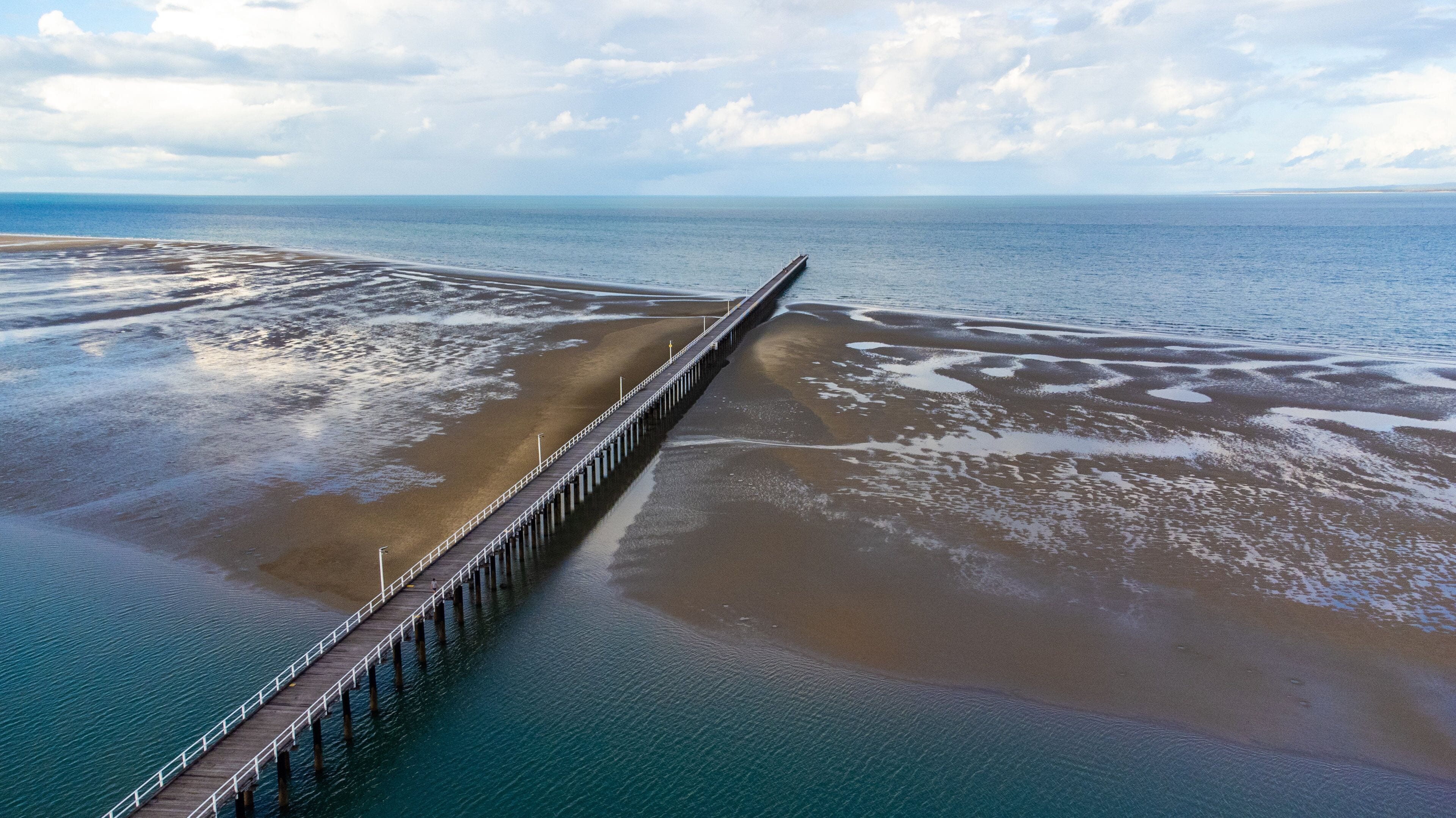 A long pier extending into a calm sea with a sandy beach on one side.