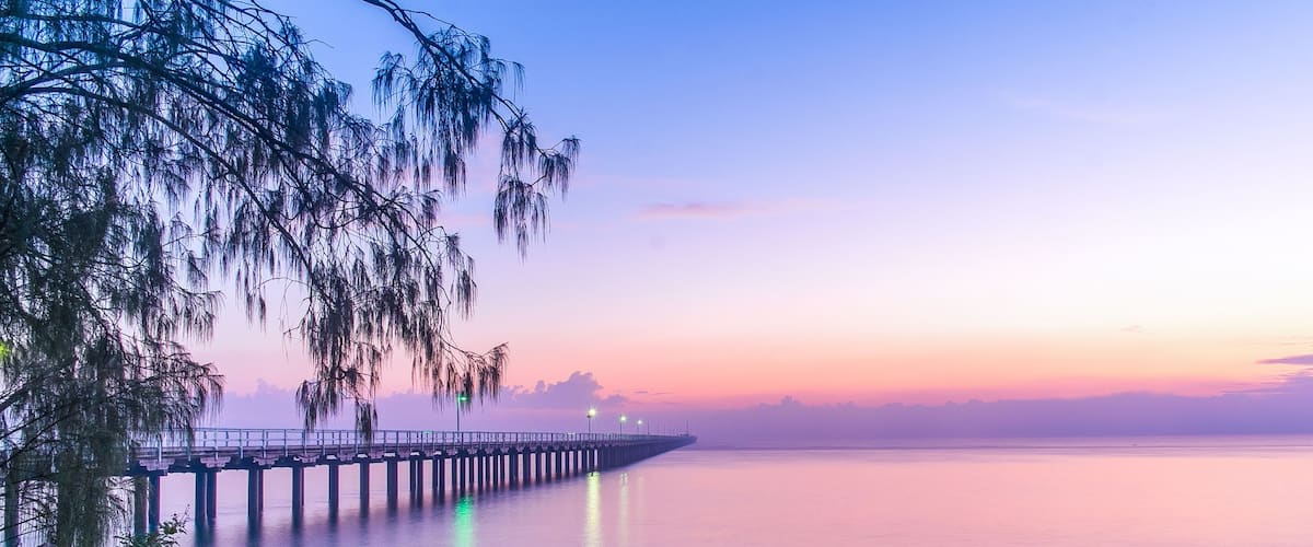 A pier extending into calm waters at sunset with a rocky foreground and trees.