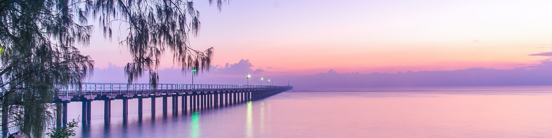 Un muelle se adentra en aguas tranquilas al atardecer, con un primer plano rocoso y árboles.