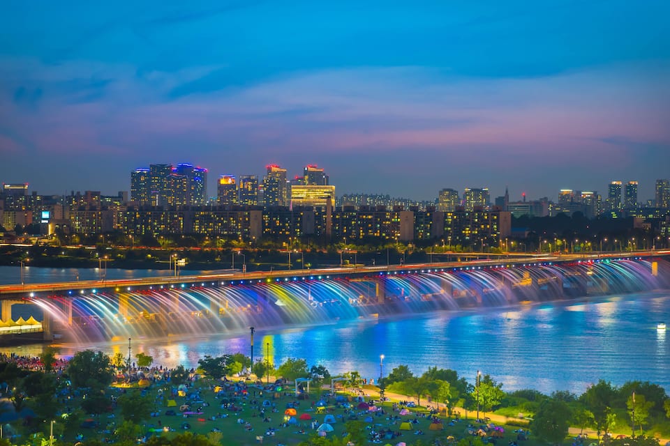 A city skyline with illuminated buildings, a waterfall over a river, and a bridge with light trails.