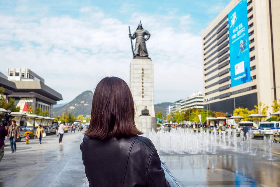 A woman in a black leather jacket facing a statue in a public square with fountains and modern buildings in the background.