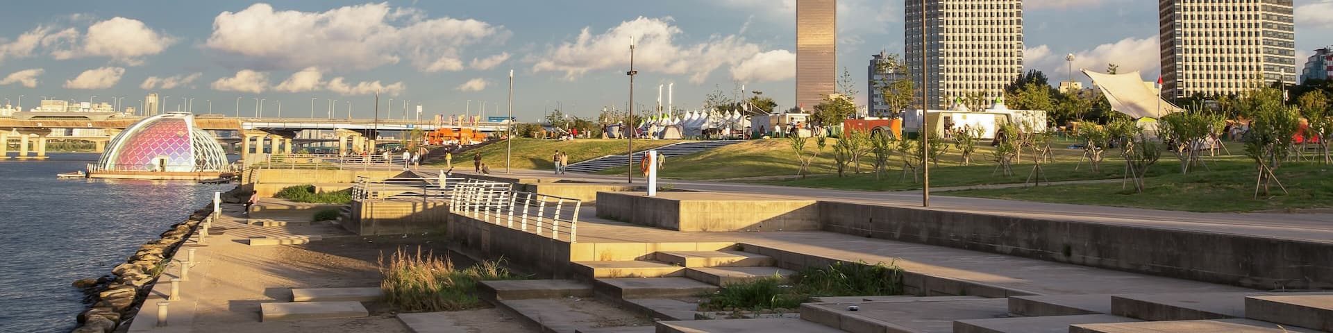 A riverside walkway with steps, a bridge, and two tall buildings in the background.