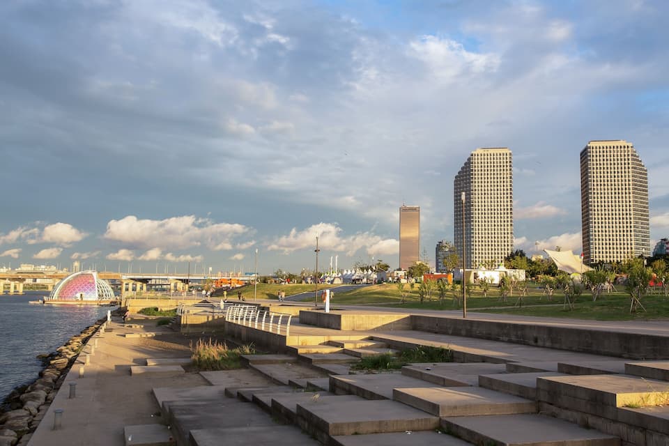 A riverside walkway with steps, a bridge, and two tall buildings in the background.