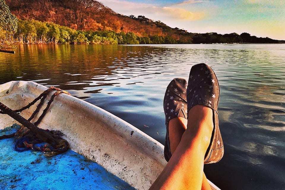 Um barco com uma pessoa relaxando, um lago calmo e uma encosta arborizada ao fundo.
