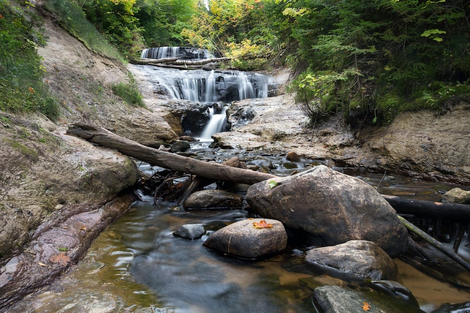 A waterfall cascading over rocks in a forested area with a fallen tree in the foreground.