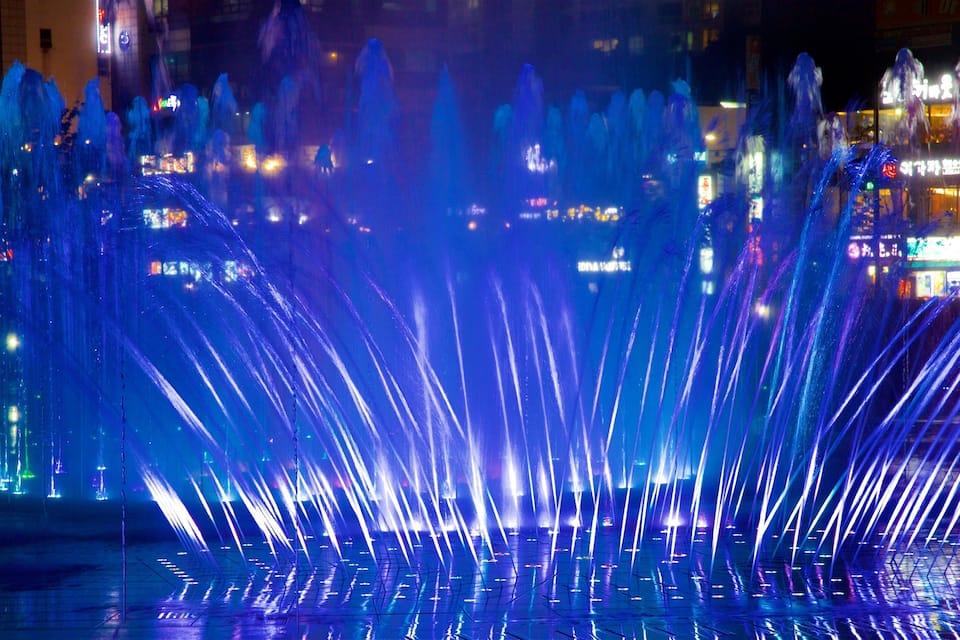 A nighttime fountain display with water jets illuminated in blue and white lights.