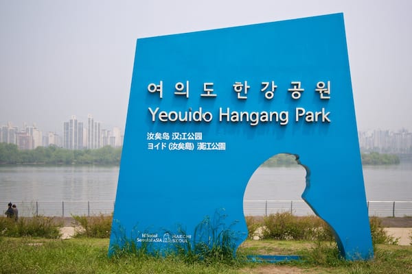 A blue sign with Korean text for Yeouido Hangang Park, a river, and a city skyline in the background.