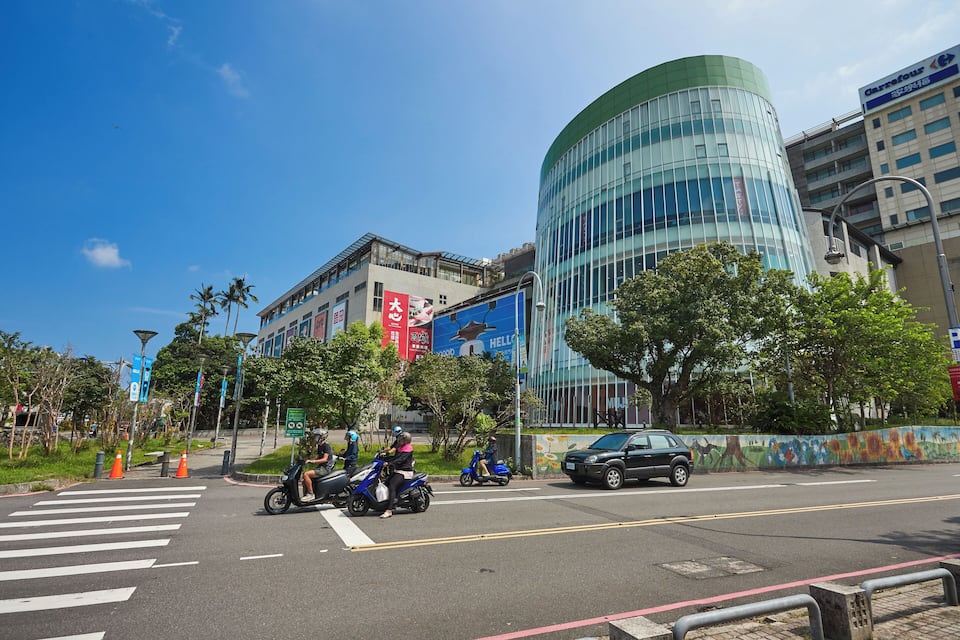 Rua movimentada na cidade, com motociclistas, um carro e edifícios modernos.