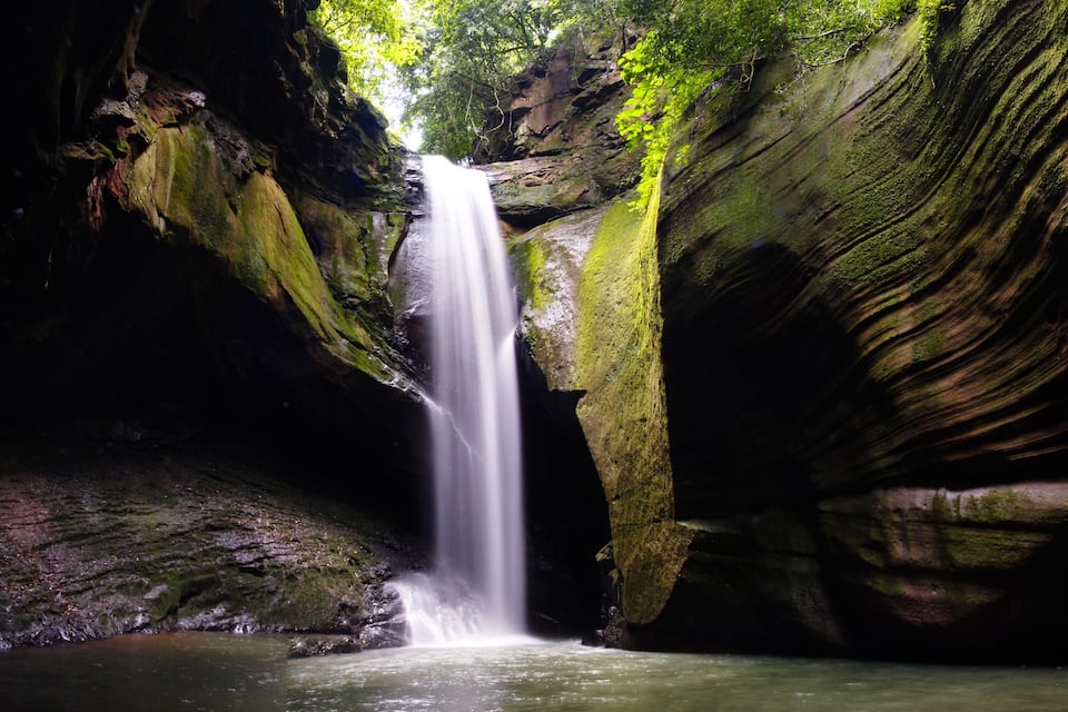 Ein Wasserfall in einer Höhle mit moosbewachsenen Steinen und einem Becken am Fuße.