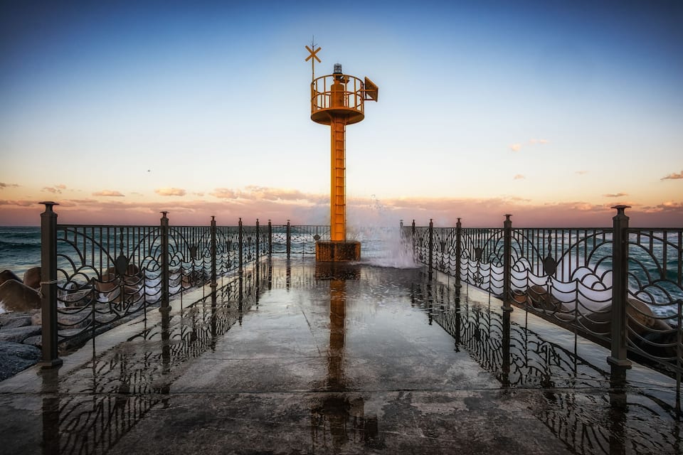 A coastal lighthouse with a metal railing and boats in the background.