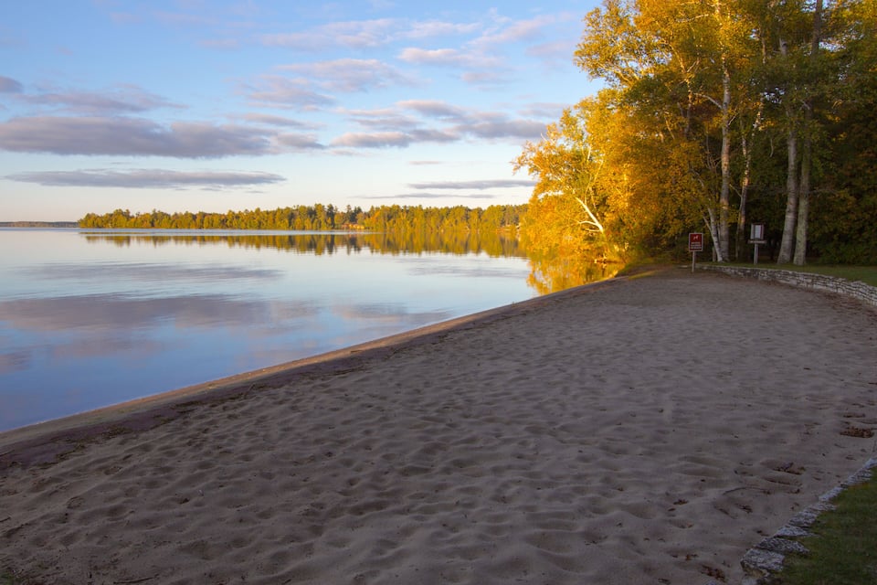 A calm lake with a sandy beach, autumn trees, and a clear sky.