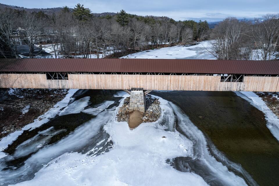 A wooden covered bridge over a partially frozen river with snow on the banks and trees in the background.