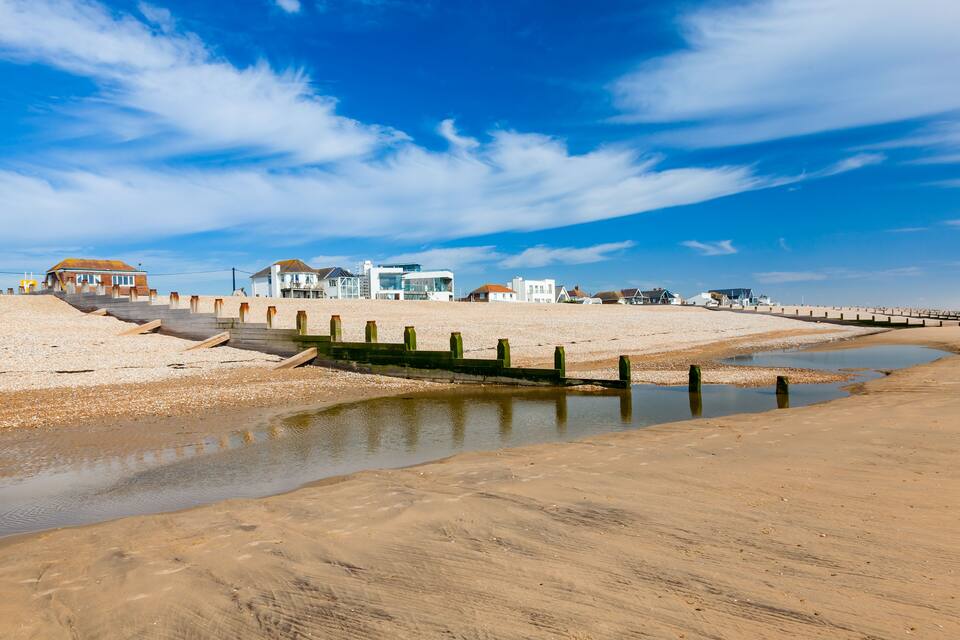 A beach with a wooden walkway leading to houses, a sandy shore, and a clear blue sky.
