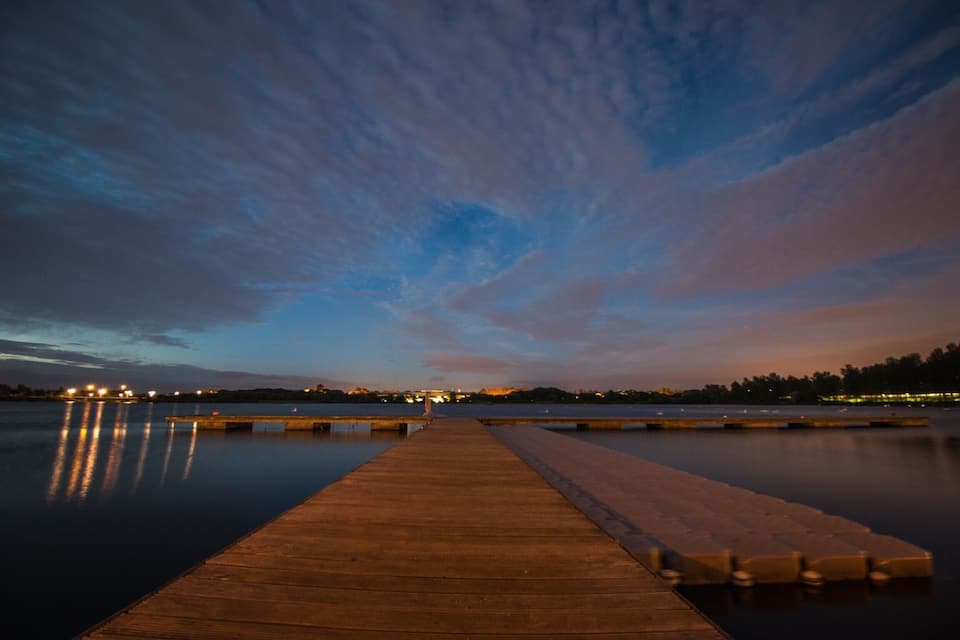 Une jetée en bois s’étendant dans des eaux calmes au crépuscule, avec la silhouette d’une ville en arrière-plan.