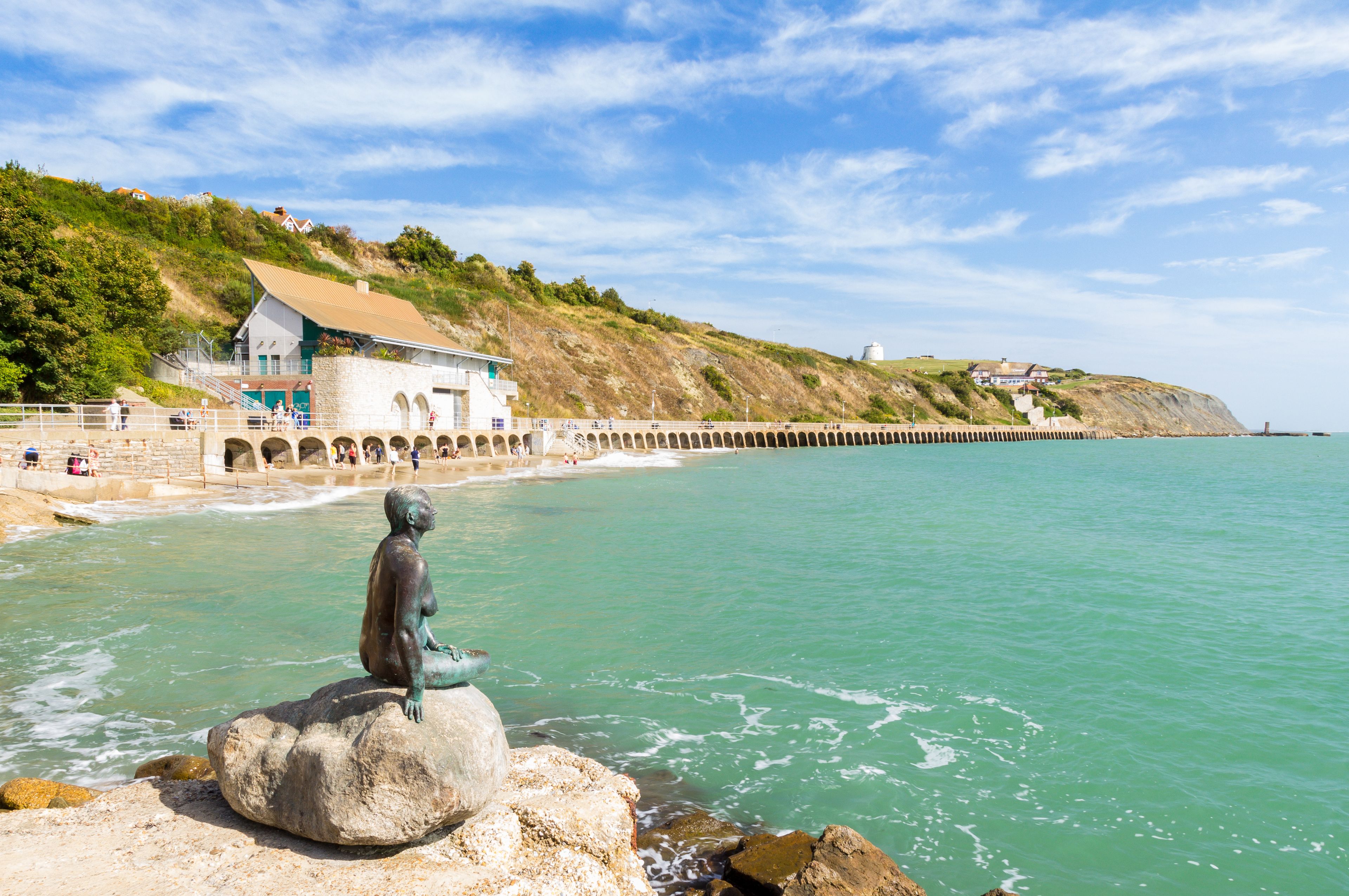 Escena costera con una estatua sobre un peñasco, un muelle que se adentra en el mar y una colina con edificios.