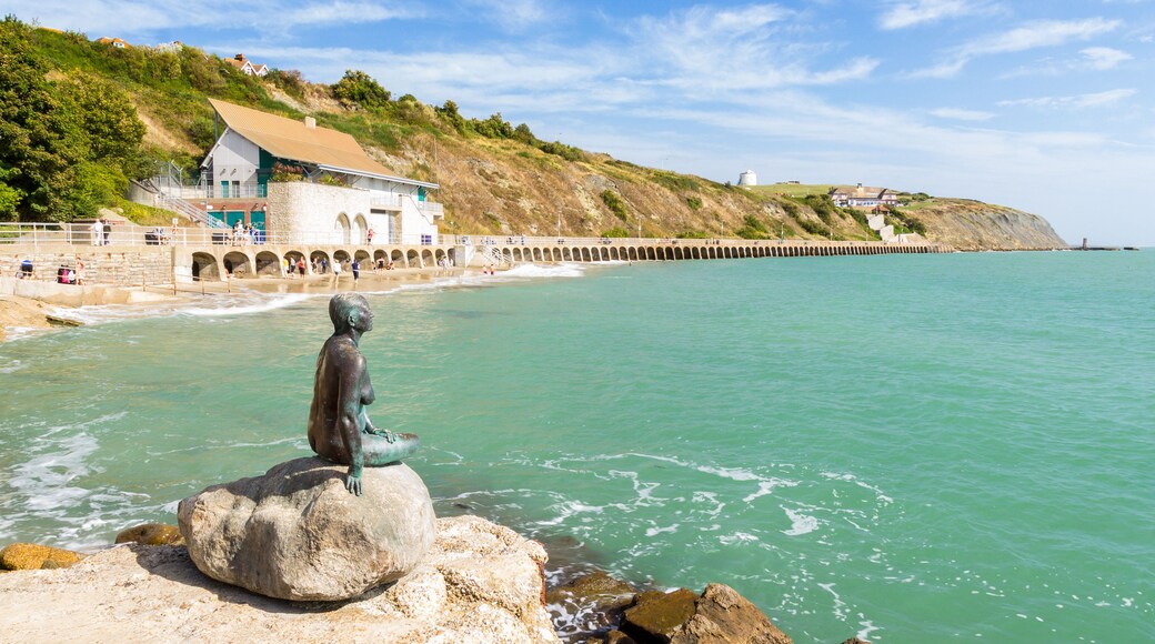 Escena costera con una estatua sobre un peñasco, un muelle que se adentra en el mar y una colina con edificios.