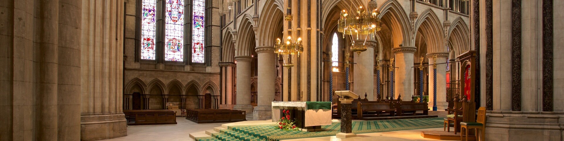 Interior imponente de uma catedral, com altos tetos abobadados, vitrais coloridos e lustres ricamente decorados.