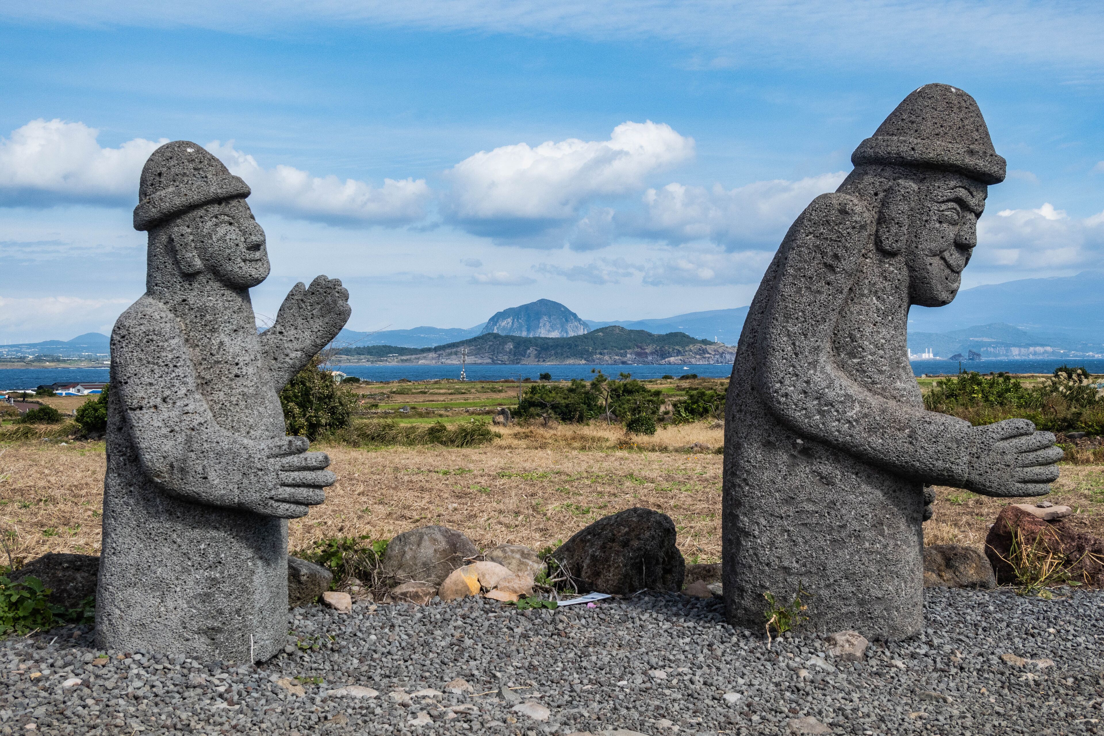 Two stone statues in a natural landscape with a distant mountain and ocean view.