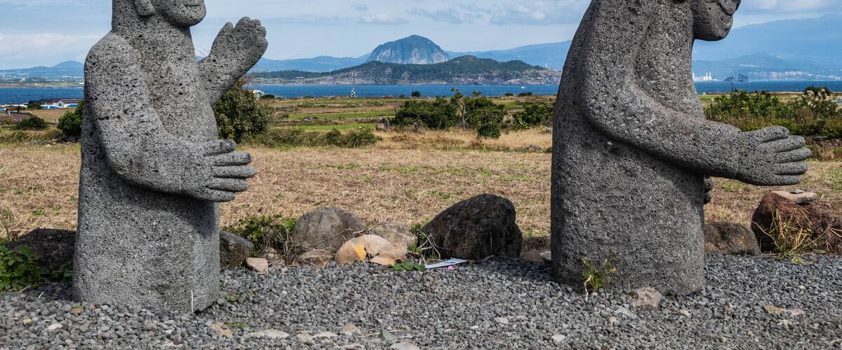 Two stone statues in a natural landscape with a distant mountain and ocean view.