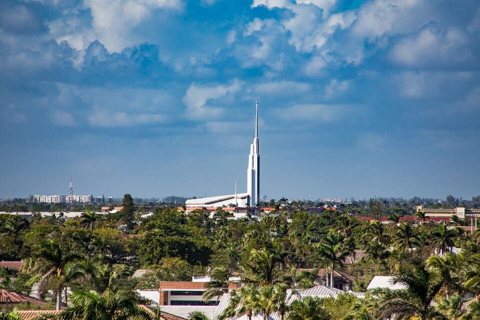 Un rascacielos alto y blanco con una aguja, rodeado de palmeras y edificios bajo un cielo parcialmente nublado.