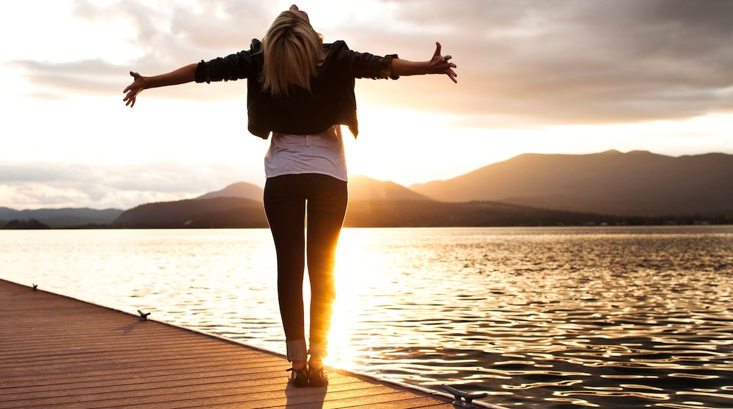 A beautiful young woman looking into the distance holds her arms out embracing the sunset over a lake in Idaho.
