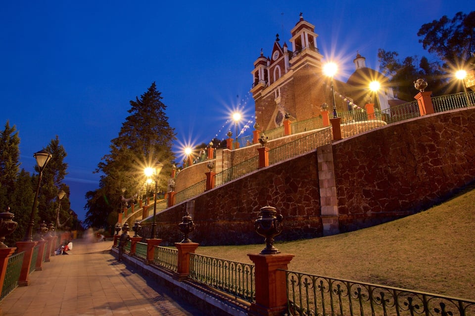 Escadaria de pedra que leva a uma igreja com torre de sino, cercada por árvores e postes de luz.