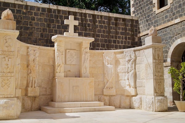 A stone monument with carvings and inscriptions, surrounded by a stone wall and a building with an arched entrance.