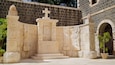 A stone monument with carvings and inscriptions, surrounded by a stone wall and a building with an arched entrance.