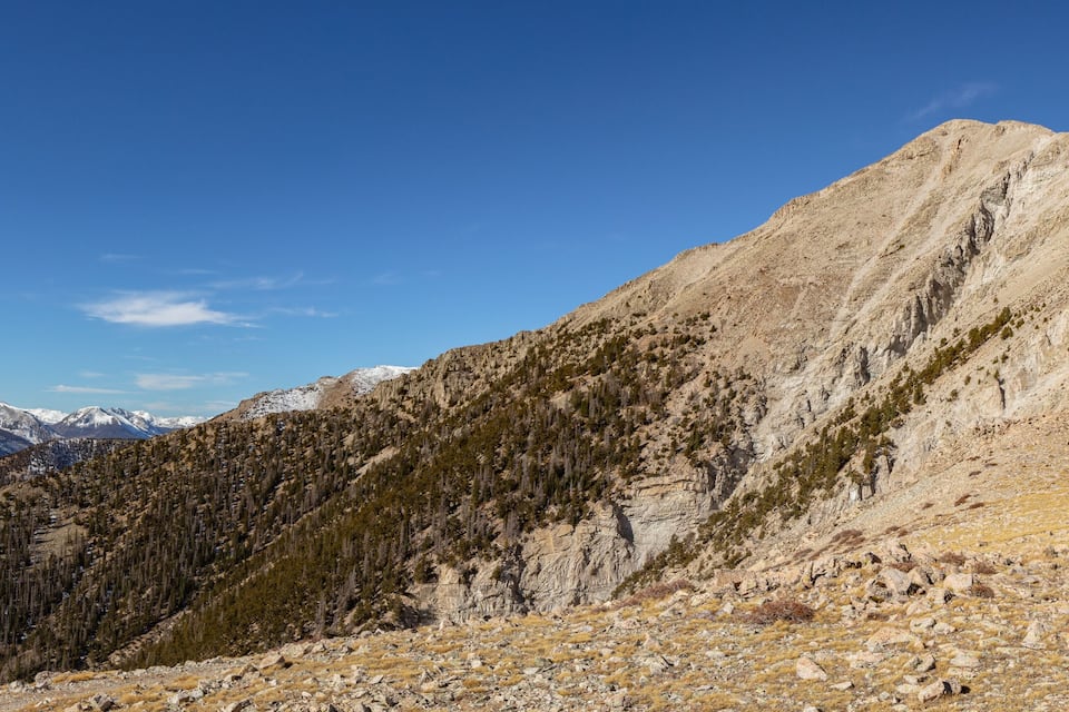 A mountain peak with a clear sky, rocky terrain, and snow-capped mountains in the background.