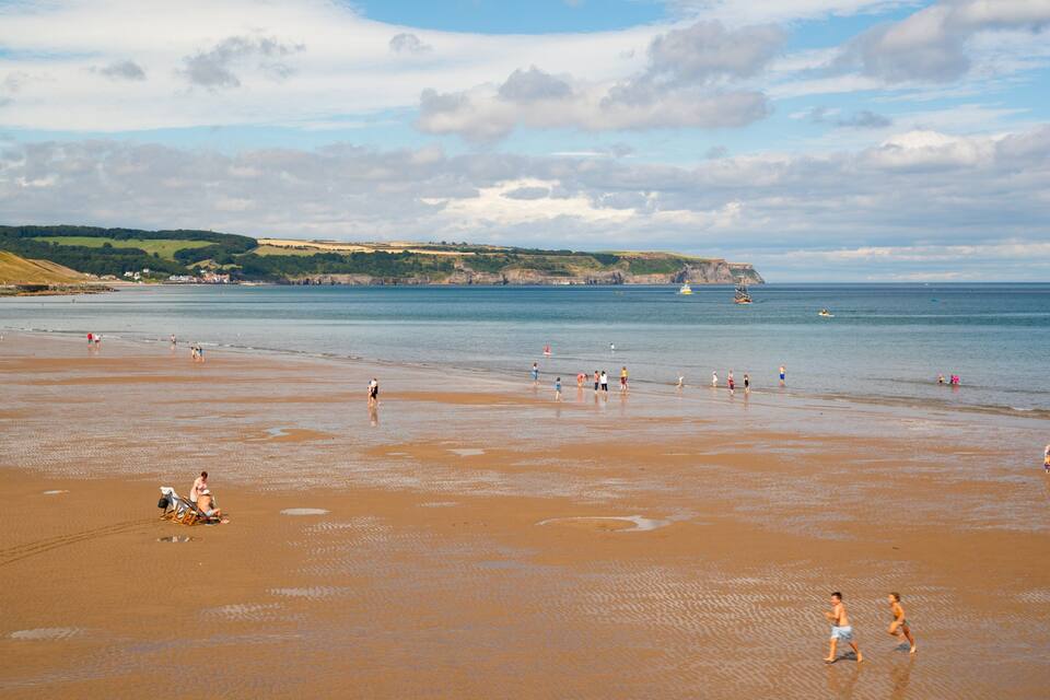 A wide sandy beach with people walking and playing, a calm sea, and hills in the background.