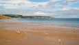 A wide sandy beach with people walking and playing, a calm sea, and hills in the background.