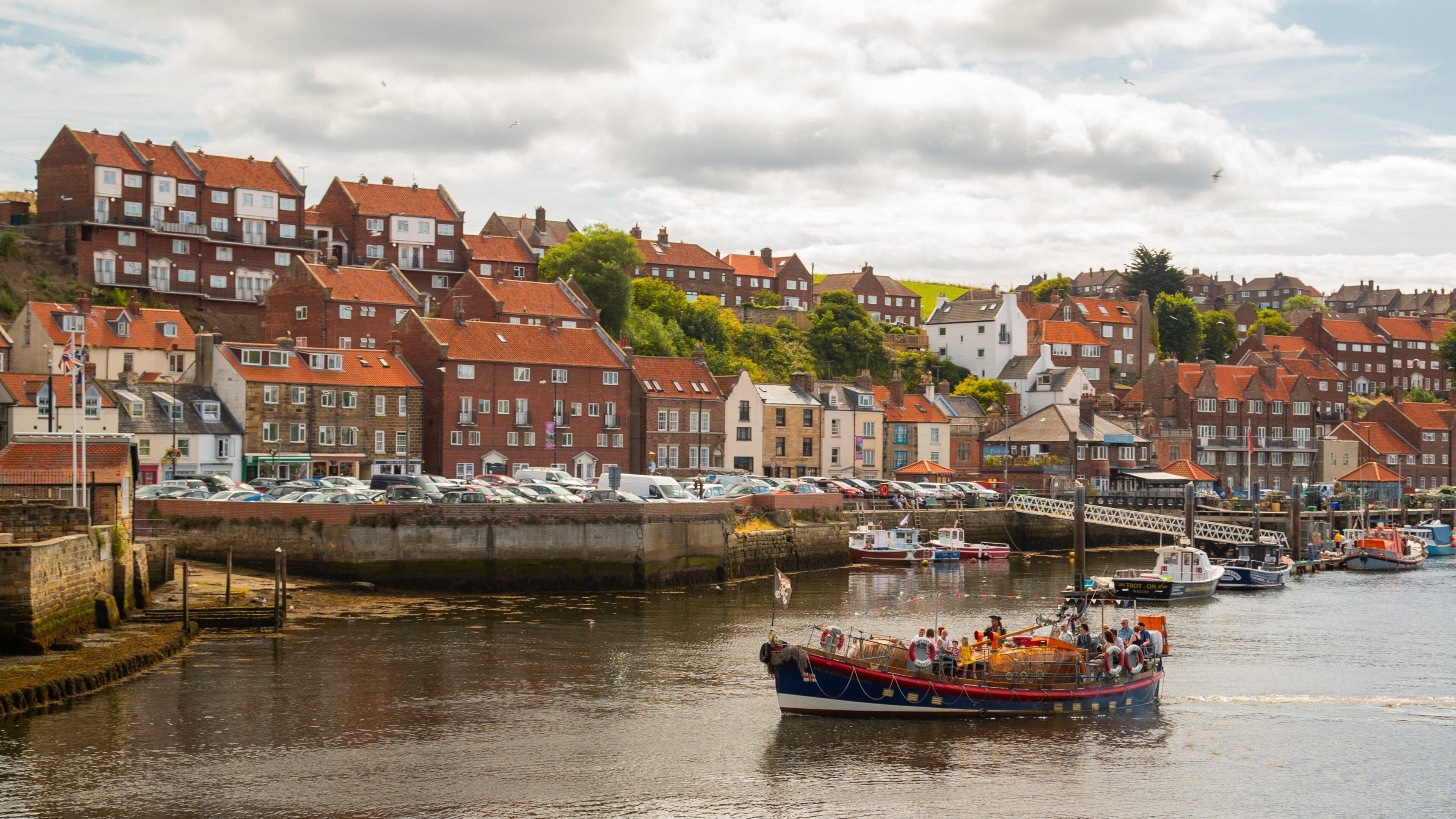 A harbor with a red-roofed boat docked at the pier, surrounded by historic buildings and a cloudy sky.