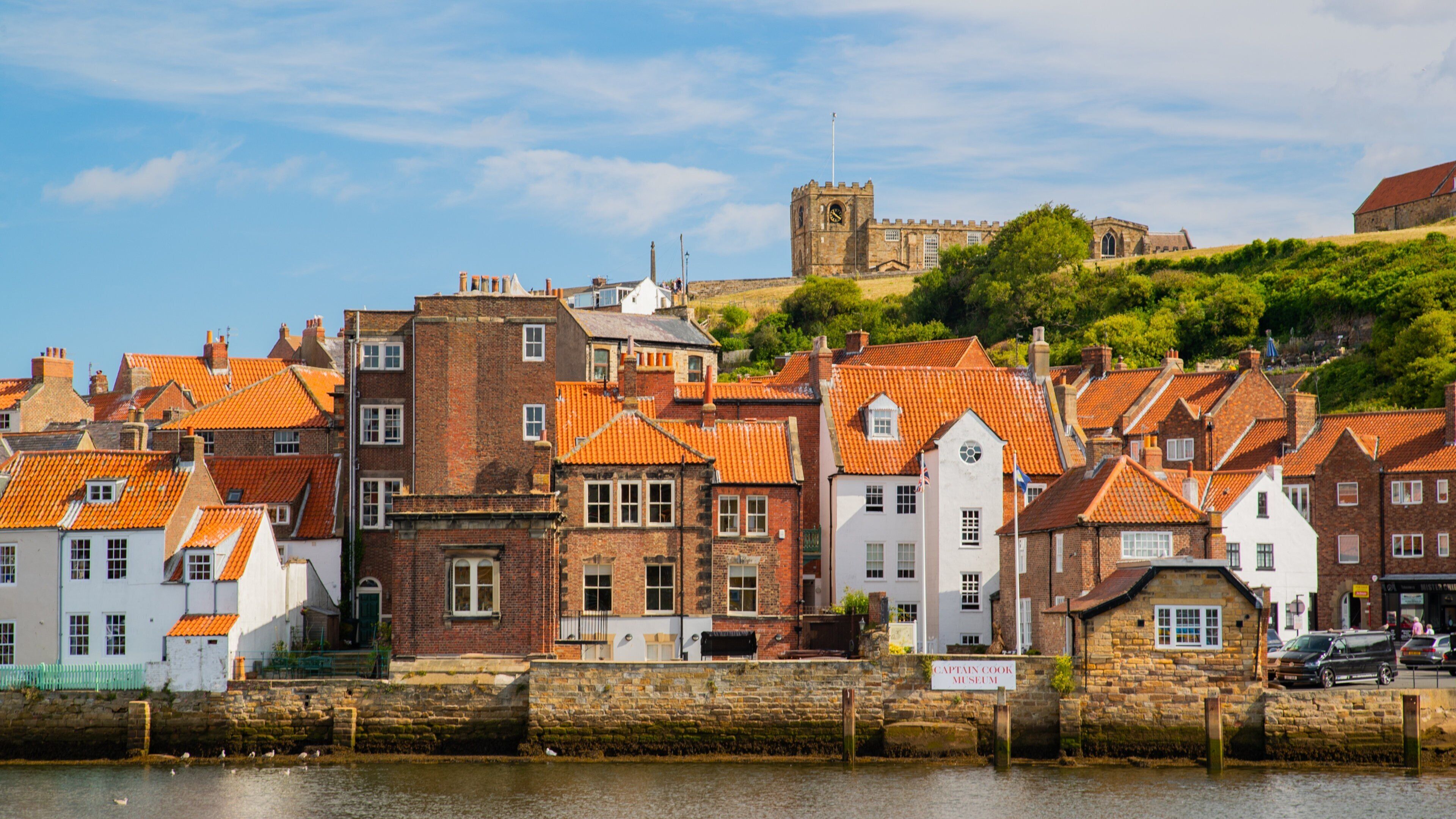 A waterfront town with red-tiled roofs, a stone wall, and a castle in the background.