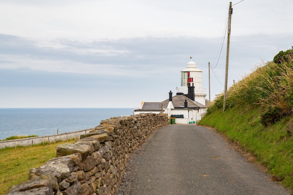 Une route côtière bordée d’un mur de pierres, un bâtiment blanc avec une cheminée rouge et, au loin, un phare.