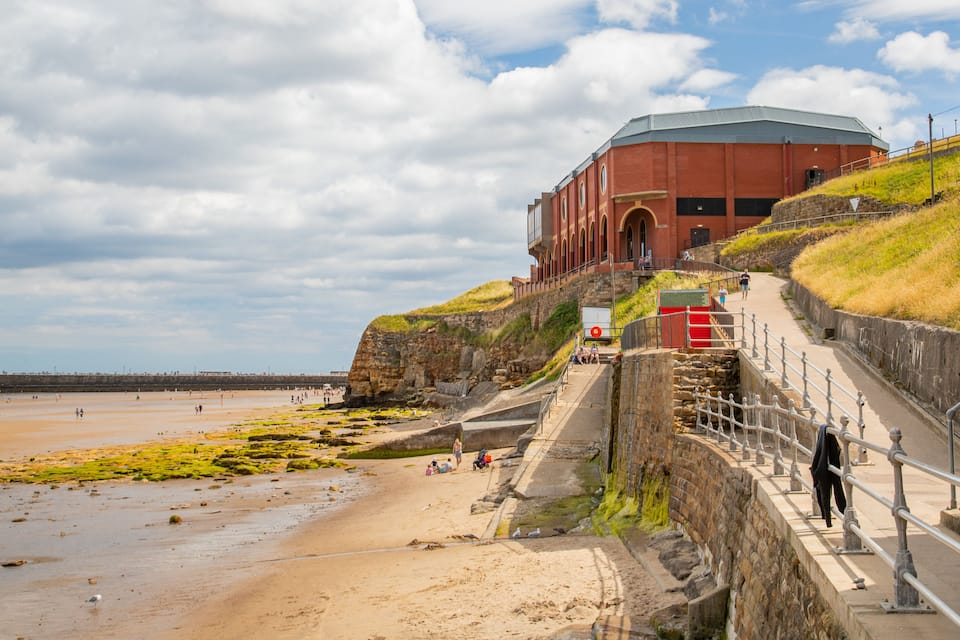 A coastal promenade with a red brick building, a beach, and a lighthouse in the distance.