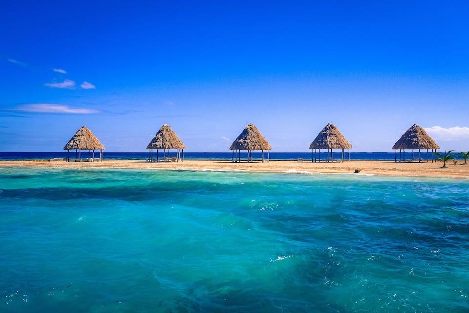 Een strand met rieten hutten en helder turquoise water.