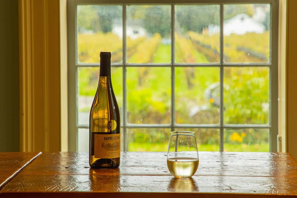 A bottle of Ravine Vineyard Chardonnay and a glass on a wooden table by a window with a vineyard view.