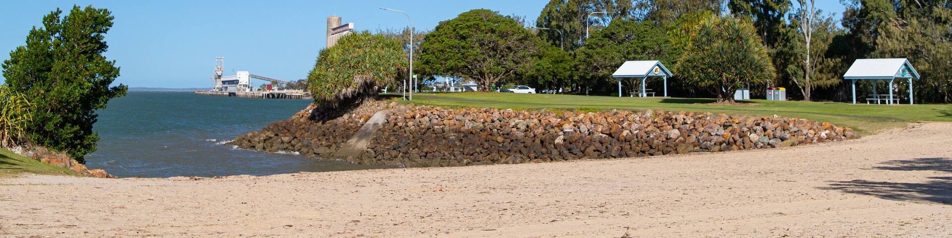 A beach with a rocky shoreline, a pier, and a park with trees and small buildings.