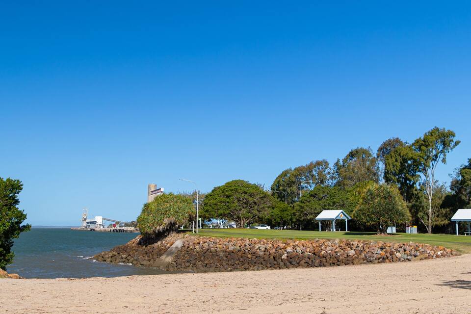 A beach with a rocky shoreline, a pier, and a park with trees and small buildings.