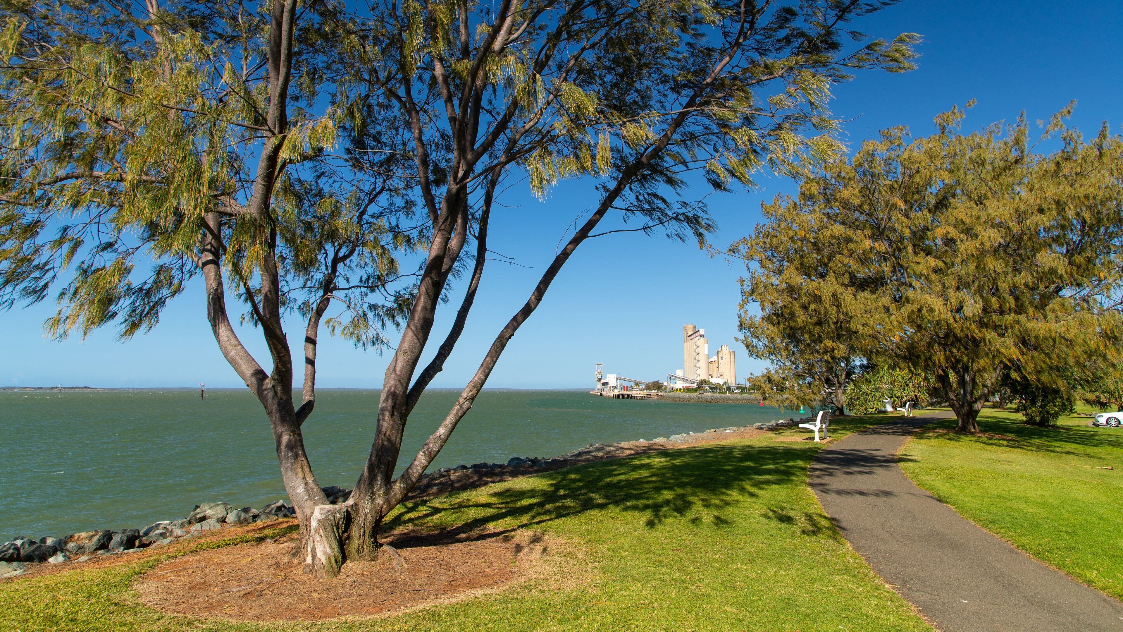 A park with a tree-lined path, a body of water, and industrial buildings in the background.