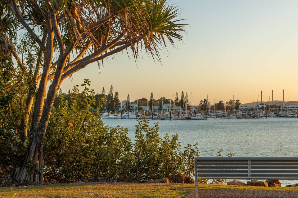 A waterfront with a bench, trees, and a marina in the background.