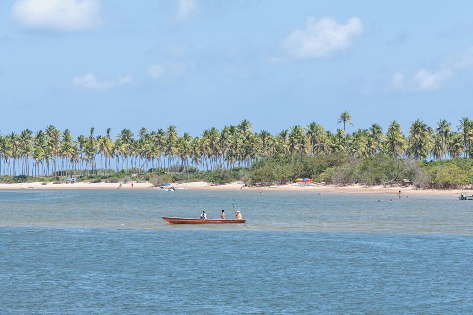 A calm sea with a wooden boat carrying two people, palm trees, and a sandy beach in the background.