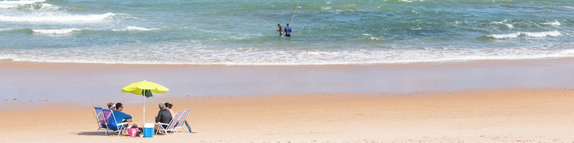 A beach with people under an umbrella, a kite surfer in the water, and waves approaching the shore.
