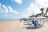 A beach with blue chairs and umbrellas, palm trees, and people walking.