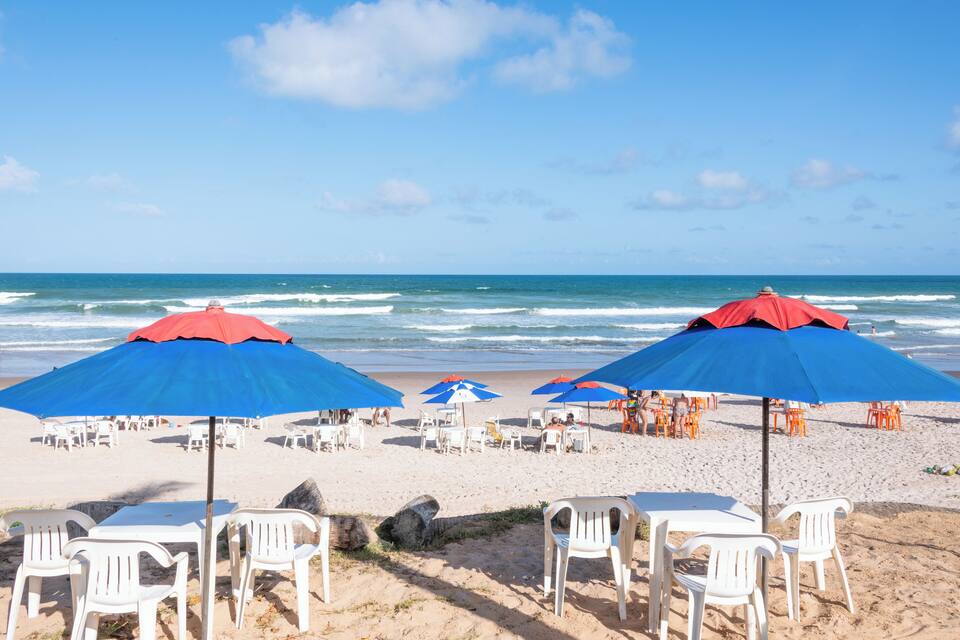 Una playa con sillas y mesas de plástico blanco, y sombrillas rojas y azules.