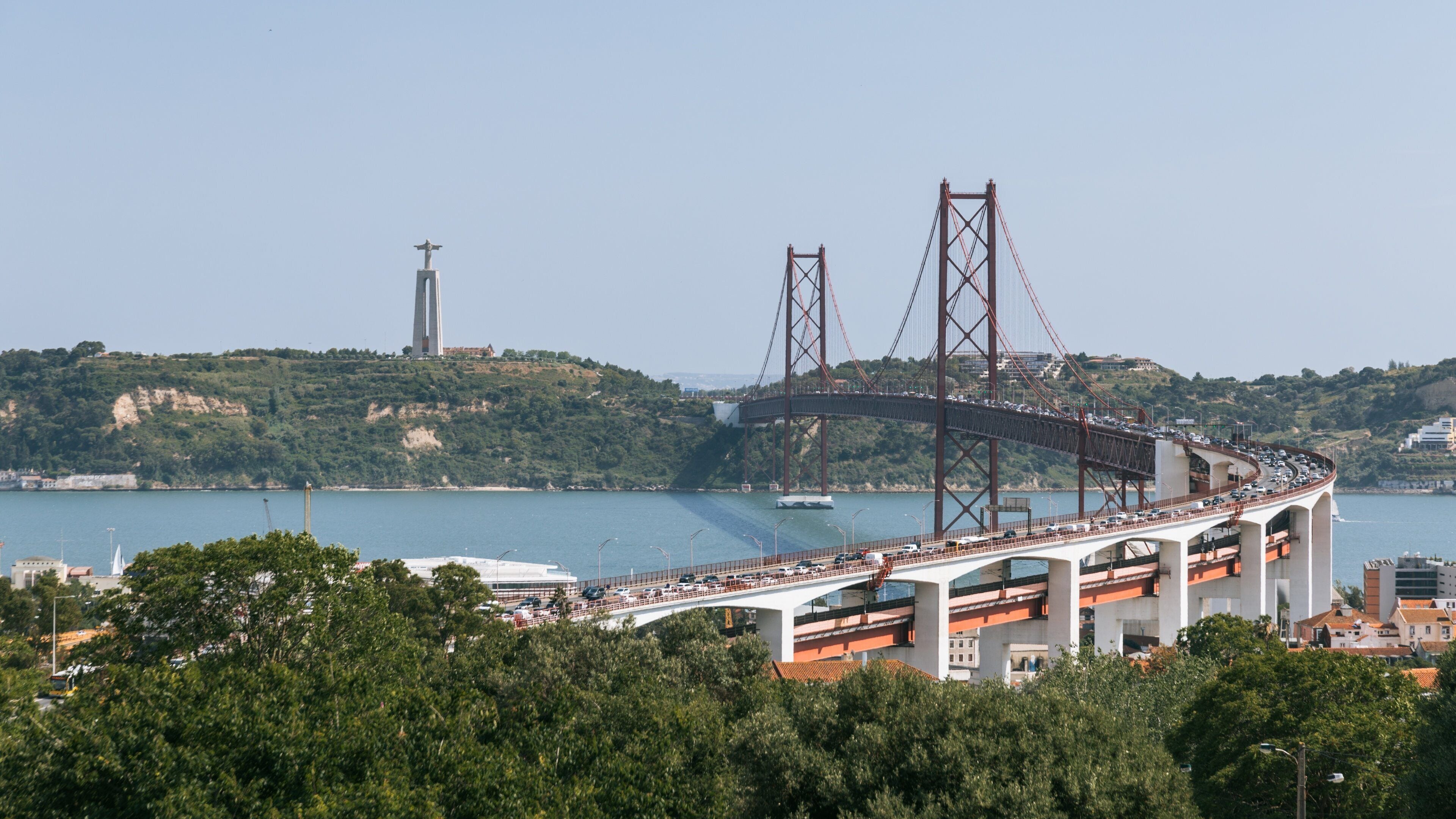 A bridge with two towers over a body of water, surrounded by greenery and buildings.