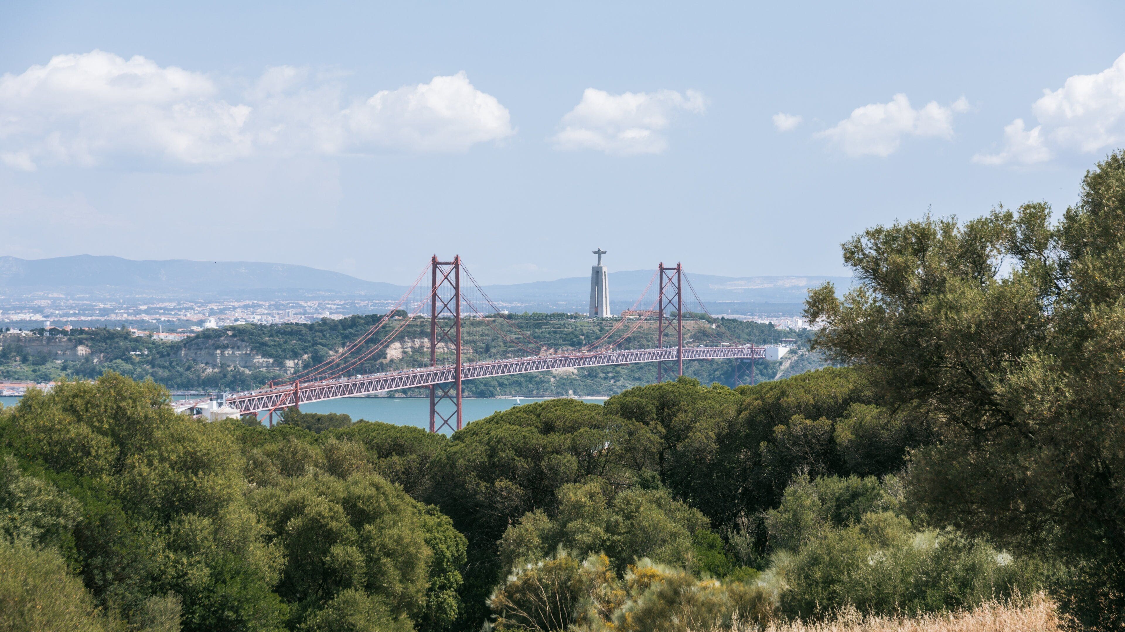 A red suspension bridge over a river, surrounded by dense green trees and hills.