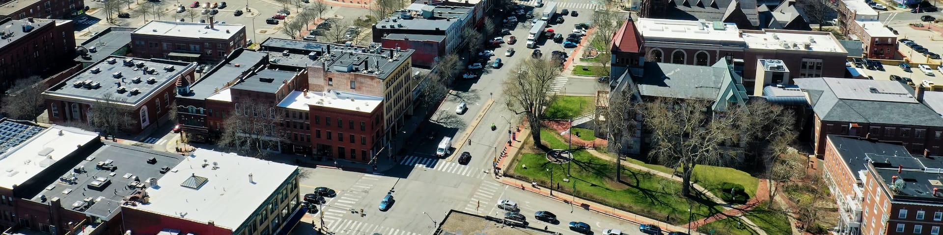 An aerial view of a town with a mix of historic and modern buildings, a central road, and surrounding hills.