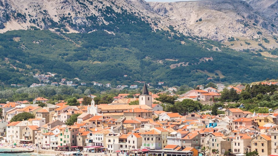 A coastal town with a marina filled with boats, surrounded by mountains and greenery.
