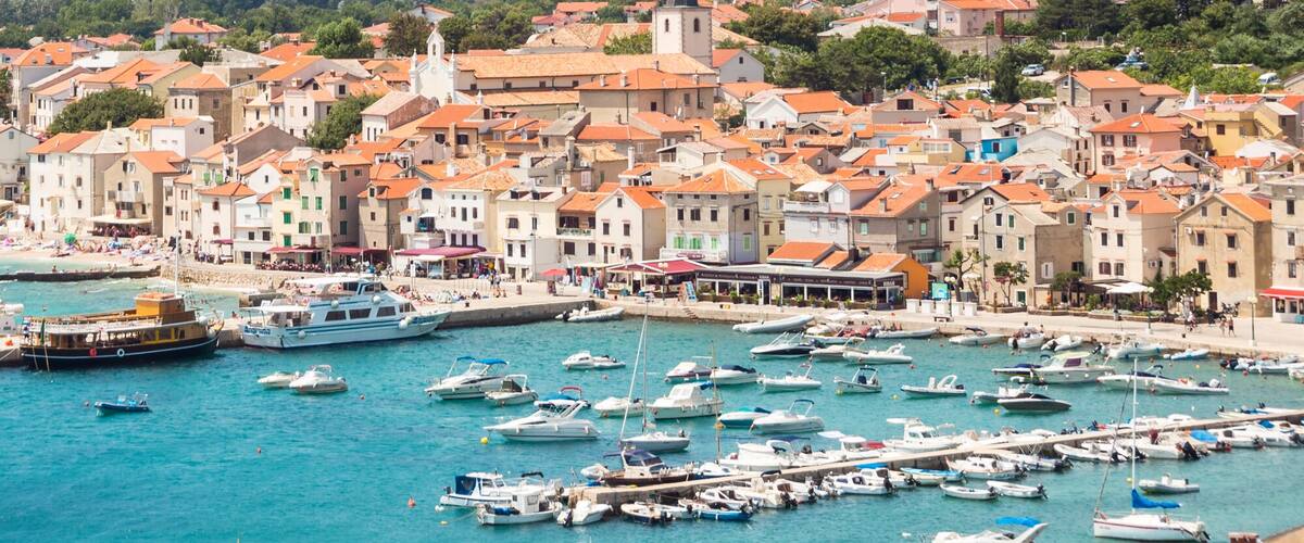 A coastal town with a marina filled with boats, surrounded by mountains and greenery.